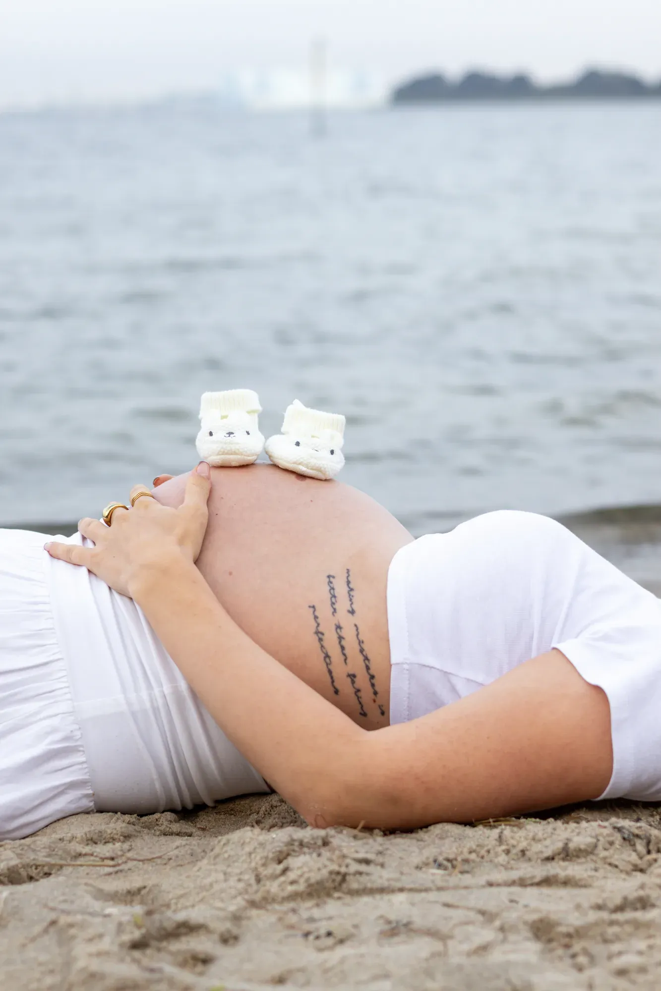Schwangere liegt im Sand am Wasser, Babyschuhe auf dem Bauch