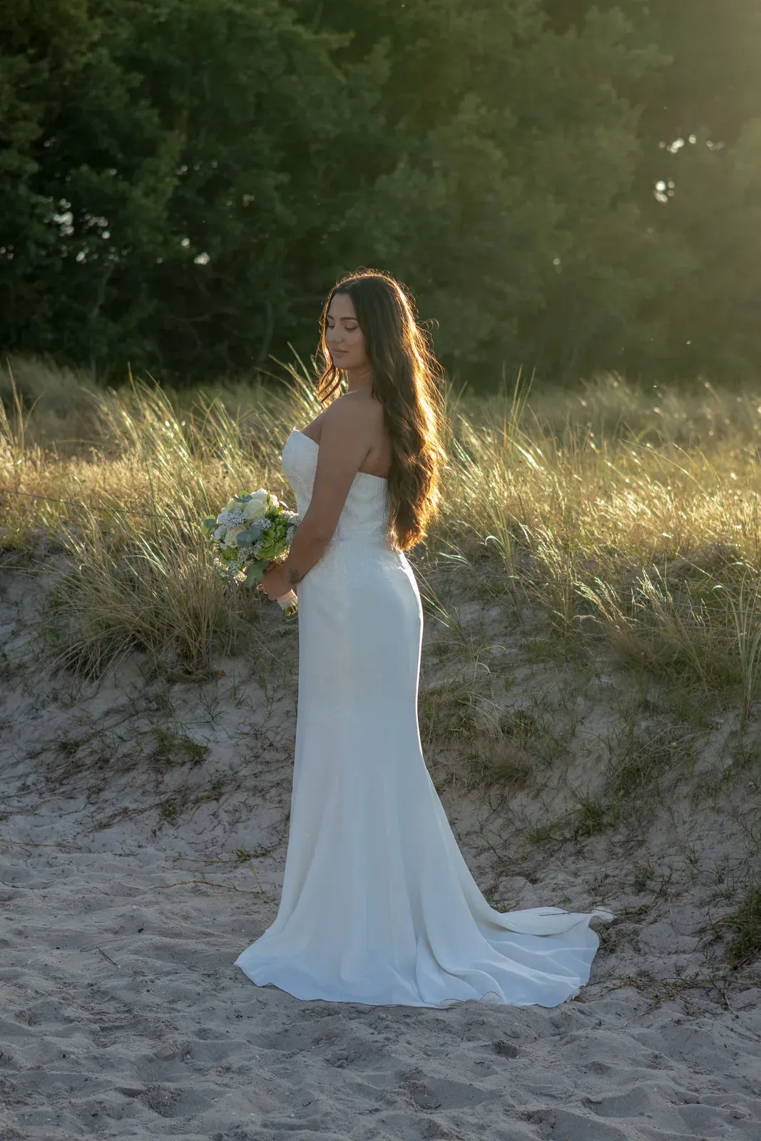 Braut mit Strauß steht in den Dünen, das Kleid im Sand