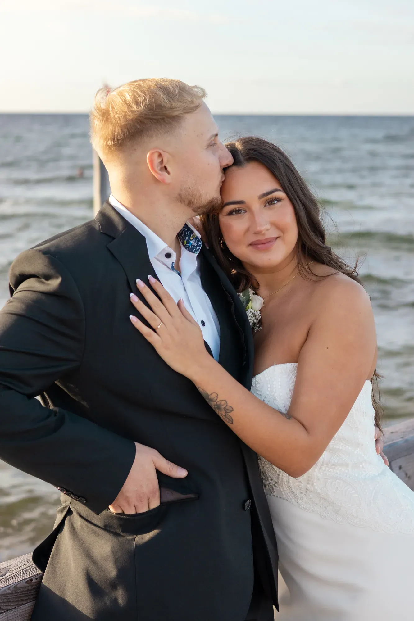 Bräutigam küsst die Braut aufs Haar, im Hintergrund das Meer