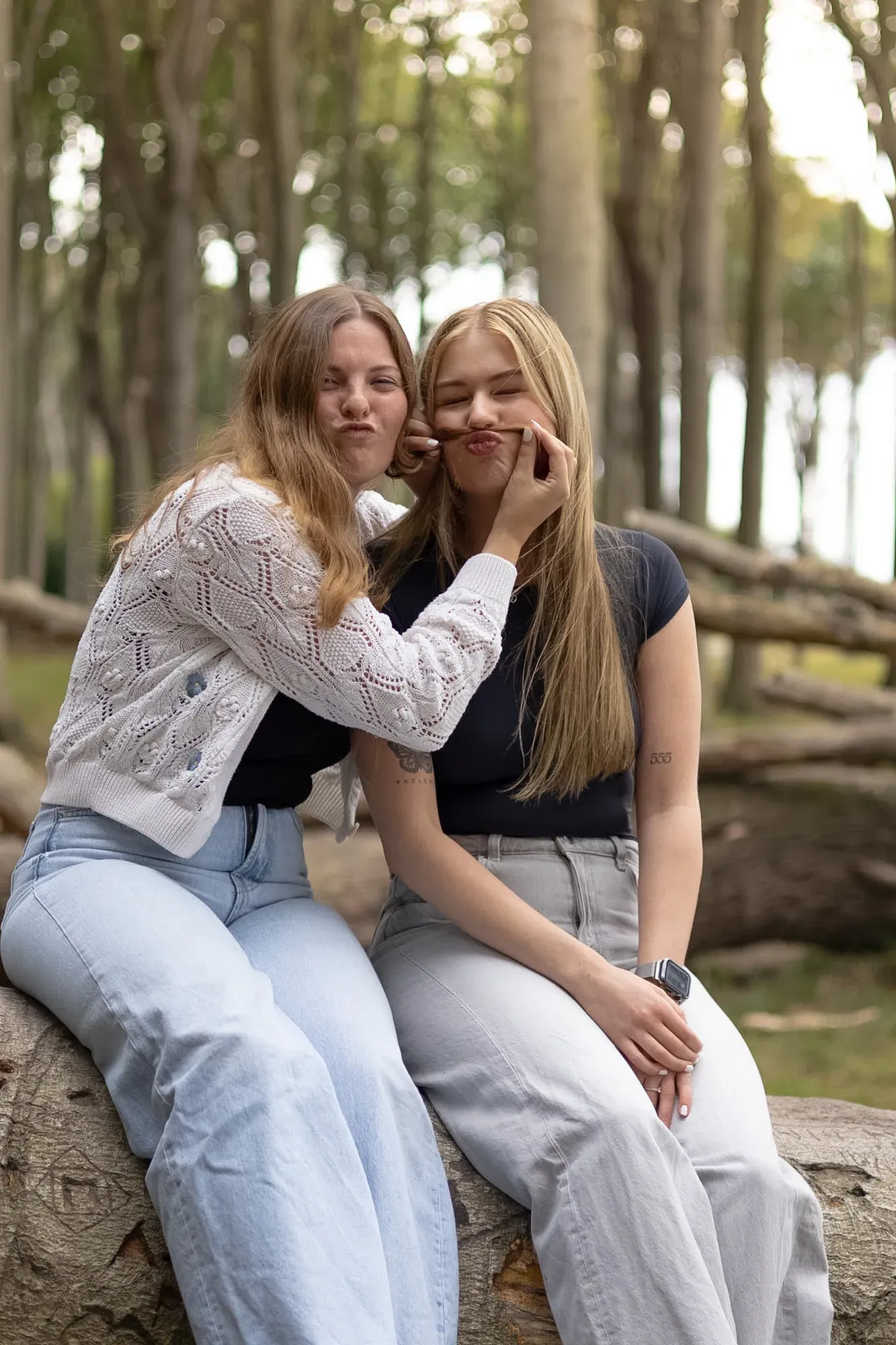 Zwei Freundinnen sitzen auf einem Baumstamm im Küstenwald, eine zieht eine Grimasse