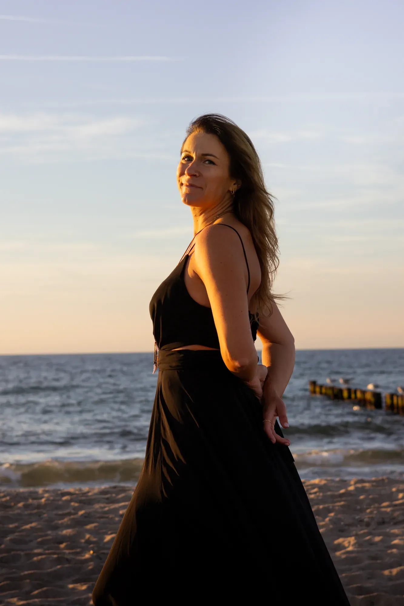 Frau im schwarzen Kleid blickt am Strand über die Schulter, dahinter die Buhnen