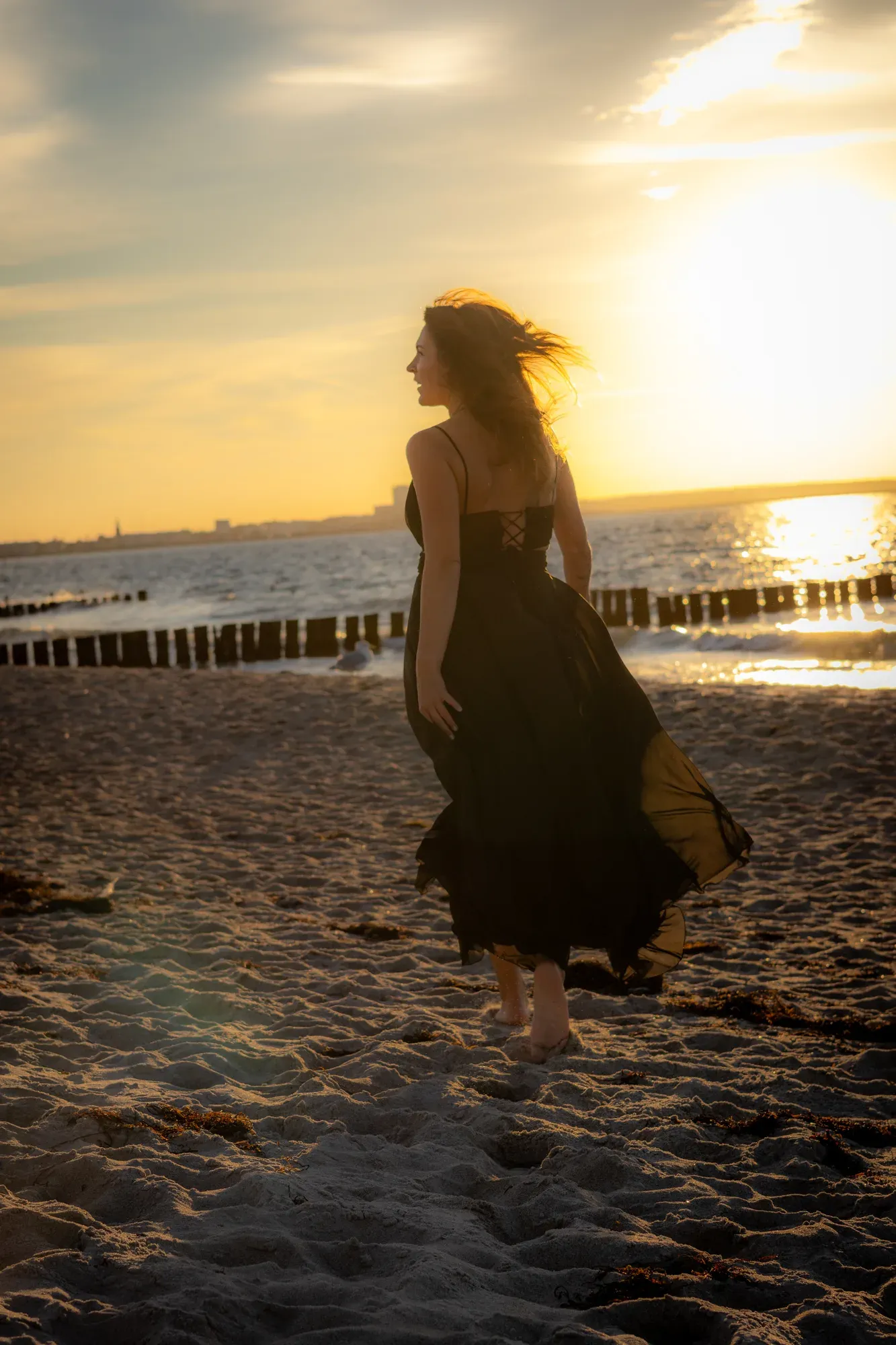 Frau im schwarzen Kleid steht am Strand, das Haar weht im Gegenlicht
